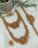 An ornate gold-toned traditional jewelry set arranged on a pale mint fabric backdrop, including a necklace, matching earrings, a maang tikka, and a long bracelet or chain. The pieces are decorated with detailed motifs, small beads, and red and green enamel accents.