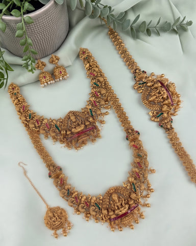 An ornate gold-toned traditional jewelry set arranged on a pale mint fabric backdrop, including a necklace, matching earrings, a maang tikka, and a long bracelet or chain. The pieces are decorated with detailed motifs, small beads, and red and green enamel accents.
