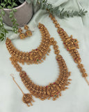 An ornate gold-toned traditional jewelry set arranged on a pale green fabric backdrop, including a necklace, matching earrings, a bracelet, a pendant, and a chain, with small red and green stone accents and decorative leaves in a pot.