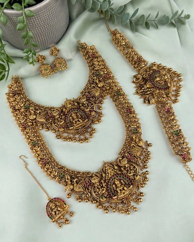 An ornate gold-toned traditional jewelry set arranged on a pale green fabric backdrop, including a necklace, matching earrings, a bracelet, and a dangling pendant. The pieces are decorated with intricate filigree, beadwork, and small multicolored stone accents.