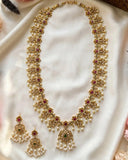 Gold-plated Indian traditional jewelry set featuring a long necklace and matching earrings, embellished with white pearls, red gemstones, and green stones, displayed on a draped white satin fabric.