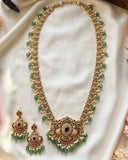 Gold-plated Indian jewelry set featuring a necklace and matching earrings with green stone beads, white pearl-like beads, and red stone accents, displayed on a white satin fabric.