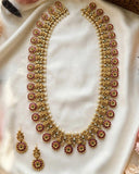 Gold-plated Indian traditional jewelry set featuring a necklace and matching earrings, embellished with red floral enameles, green accents, and white faux pearls, displayed on a white satin fabric.