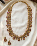 An ornate gold-toned traditional necklace set with matching earrings, decorated with red and green stone accents, displayed on a draped cream satin fabric.