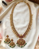 Gold-plated Indian ethnic jewelry set featuring a double-strand necklace with a large round pendant and matching drop earrings, embellished with red stones, green beads, and white pearls, displayed on a draped white fabric background.