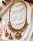 An ornate gold-toned traditional necklace set with matching earrings, decorated with green and red stone accents and white pearl-like beads, arranged on a white satin fabric with soft pink flowers around the edges.