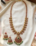 Gold-toned traditional Indian jewelry set featuring a necklace with a large pendant and matching earrings, embellished with red stones, green stones, and white faux pearls, displayed on a draped white fabric background.