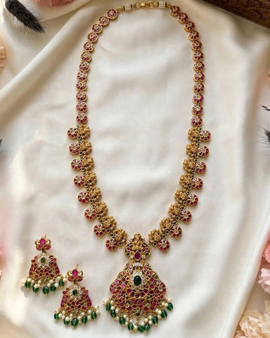 Gold-toned traditional Indian jewelry set featuring a necklace with a large pendant and matching earrings, embellished with red stones, green stones, and white faux pearls, displayed on a draped white fabric background.