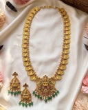Gold-toned traditional necklace and matching earrings laid on white satin fabric, decorated with coin-like links, a central deity pendant, and green and pink stone accents.
