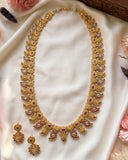 An ornate gold-toned traditional necklace with matching drop earrings, decorated with pink, red, and green stones, displayed on a draped white fabric background.