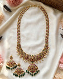 An ornate Indian-style jewelry set featuring a gold-plated necklace, matching earrings, and a pendant, embellished with red stones, green beads, and white pearls, displayed on a white satin fabric with decorative flowers.