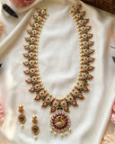 Gold-plated Indian traditional jewelry set featuring a necklace and matching earrings, embellished with white pearls, red stones, and green stones, displayed on a white satin fabric.