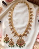 An ornate Indian-style gold necklace and matching earrings set, decorated with white pearls, green stones, and pink/red enamel, displayed on a white satin fabric with decorative flowers.