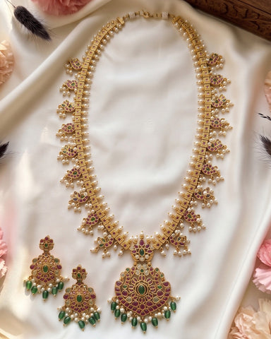 An ornate Indian-style gold necklace and matching earrings set, decorated with white pearls, green stones, and pink/red enamel, displayed on a white satin fabric with decorative flowers.