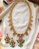 Gold-toned traditional Indian jewelry set featuring a necklace and matching earrings, embellished with white pearls, red stones, and green beads, displayed on a draped white satin fabric with pink flowers.