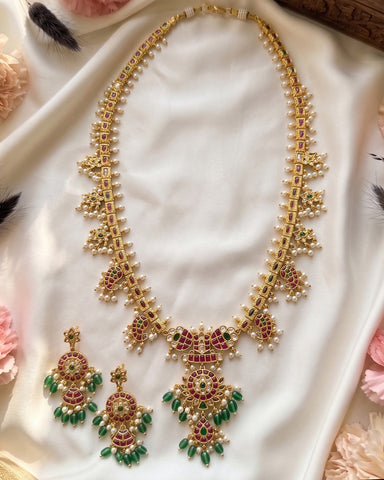 Gold-toned traditional Indian jewelry set featuring a necklace and matching earrings, embellished with white pearls, red stones, and green beads, displayed on a draped white satin fabric with pink flowers.