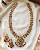 Gold-toned traditional Indian jewelry set featuring a necklace with a large central pendant and matching drop earrings, decorated with red and green stones and white pearl-like beads, displayed on a white satin fabric.