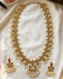 Gold-toned traditional necklace and matching drop earrings adorned with small white pearls, displayed on a soft white fabric background.