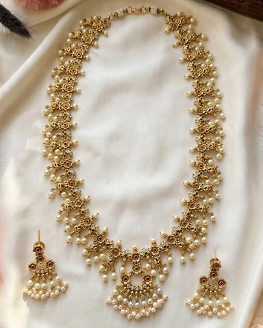 Gold-toned traditional necklace and matching drop earrings adorned with small white pearls, displayed on a soft white fabric background.
