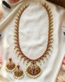 Gold-plated Indian jewelry set featuring a necklace and matching earrings embellished with red and green stones, pearls, and gold, displayed on a draped white satin fabric.