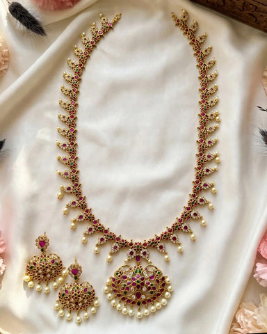 Gold-plated Indian jewelry set featuring a necklace and matching earrings embellished with red and green stones, pearls, and gold, displayed on a draped white satin fabric.