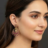A close-up of a woman wearing a large gold-toned traditional Indian chandelier earring adorned with deep pinkish-red stones and small green stones, set against a dark gray background.