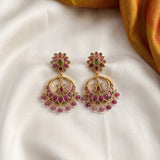 A pair of gold-toned Indian chandelier earrings with pink and green stones, displayed on a white fabric background with a gold fabric corner.