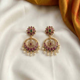 A pair of gold-toned Indian-style dangle earrings with green and pink enamel floral studs and pearl drops, displayed on a white satin fabric with a gold fabric accent in the corner.