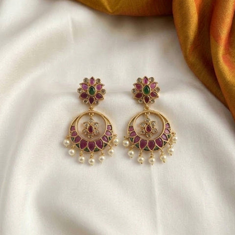 A pair of gold-toned Indian-style dangle earrings with green and pink enamel floral studs and pearl drops, displayed on a white satin fabric with a gold fabric accent in the corner.