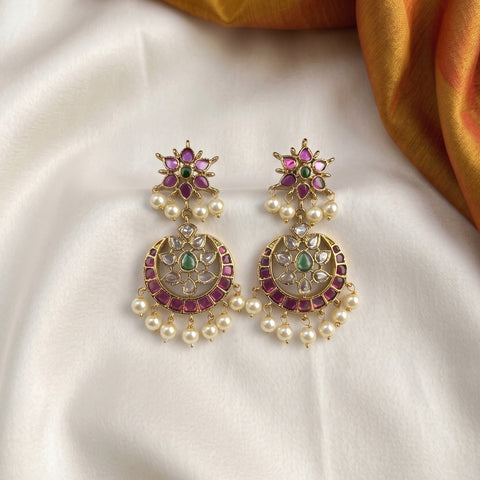 A pair of ornate gold-toned Indian jhumka earrings decorated with pink stones, green stones, and white faux pearls, displayed on a white satin fabric with a gold fabric corner in the background.