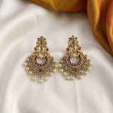 A pair of ornate gold-toned chandelier earrings with peacock motifs, white faux pearls, and green and purple stones, displayed on a white satin fabric with a gold fabric corner.