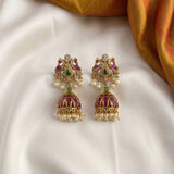 A pair of ornate gold-toned Indian jhumka earrings with purple enamel, green stone accents, and white pearl bead drops, displayed on a draped white fabric with a gold fabric corner in the background.