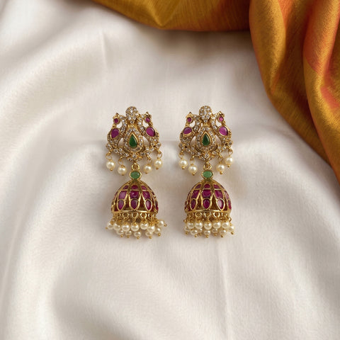 A pair of ornate gold-toned Indian jhumka earrings with purple enamel, green stone accents, and white pearl bead drops, displayed on a draped white fabric with a gold fabric corner in the background.