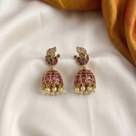 A pair of gold-plated Indian jhumka earrings decorated with pinkish-red stones, green stones, clear crystals, and dangling white pearls, displayed on a white satin fabric with a gold fabric accent in the top right corner.