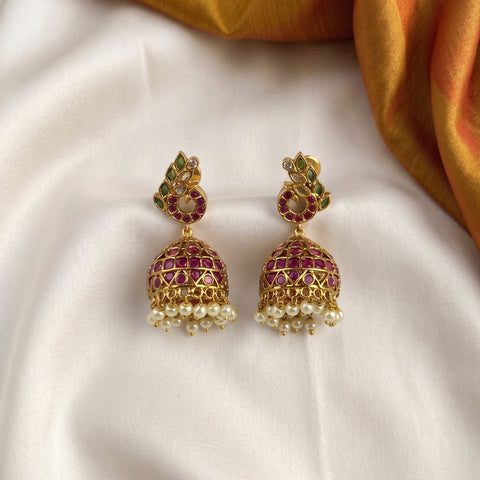 A pair of gold-plated Indian jhumka earrings with red stone embellishments, green stone accents, and dangling pearl beads, displayed on a white satin fabric with a gold fabric corner.