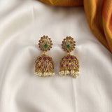 A pair of ornate gold-toned jhumka earrings with pinkish-red stones, a central green stone, and dangling white pearls, displayed on a white satin fabric with a gold fabric corner.