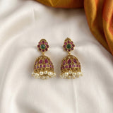 A pair of traditional Indian gold jhumka earrings with green stone floral tops, pink stone halos, and dangling white pearl chains, displayed on a white satin fabric with a gold fabric accent in the corner.