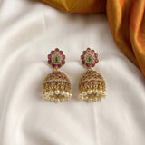A pair of ornate gold-toned Indian jhumka earrings with pink gemstone accents, a central green stone, and dangling white pearl chains, displayed on a white satin fabric with a gold fabric accent in the corner.