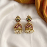 A pair of ornate gold-toned Indian jhumka earrings with pink enamel, clear stones, and dangling white pearls, displayed on a white satin fabric with a gold fabric accent in the corner.
