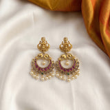 A pair of ornate gold-toned Indian chandelier earrings with pinkish-red stone accents, white pearl beads, and small green stone details, displayed on a white satin fabric with a gold fabric corner.