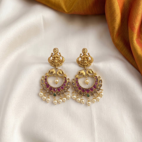 A pair of ornate gold-toned Indian chandelier earrings with pinkish-red stone accents, white pearl beads, and small green stone details, displayed on a white satin fabric with a gold fabric corner.