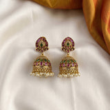 A pair of traditional Indian gold jhumka earrings with pink and green enamel detailing and white pearl bead drops, displayed on a white satin fabric with a gold fabric corner.