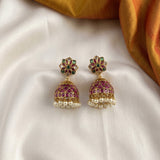 A pair of Indian jhumka earrings with gold-plated metal, pink enamel, green stone accents, and white pearl fringe, displayed on a white satin fabric with a gold fabric accent in the corner.