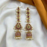A pair of gold-plated Indian jhumka earrings embellished with purple enamel, white pearls, and small green stones, displayed on a white satin fabric with a gold ribbon.