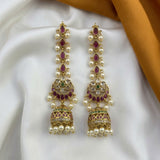 A pair of gold-plated Indian Jhumka earrings decorated with white pearls, red stones, and green accents, displayed on a white satin fabric with a gold-colored fabric drape in the background.