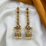 A pair of gold-plated Indian jhumka earrings decorated with white faux pearls, pinkish-red enamel, and a small green stone, displayed on a white satin fabric with a gold ribbon in the background.