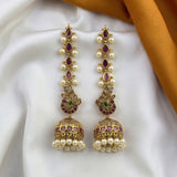 A pair of gold-plated Indian chandelier earrings with dangling pearl beads, purple enamel accents, and a small green stone detail, displayed on a draped white fabric with an orange satin corner.