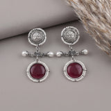 A pair of silver-toned dangle earrings with white pearl-like beads, ornate filigree tops, and oval deep red gemstones, displayed on a neutral gray surface with dried grass in the corner.