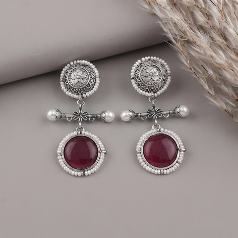 A pair of silver-toned dangle earrings with white pearl-like beads, ornate filigree tops, and oval deep red gemstones, displayed on a neutral gray surface with dried grass in the corner.