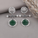 A pair of silver-toned dangle earrings with white pearl-like bead borders, green oval stones, and decorative filigree details, displayed on a neutral gray surface with dried pampas grass in the corner.